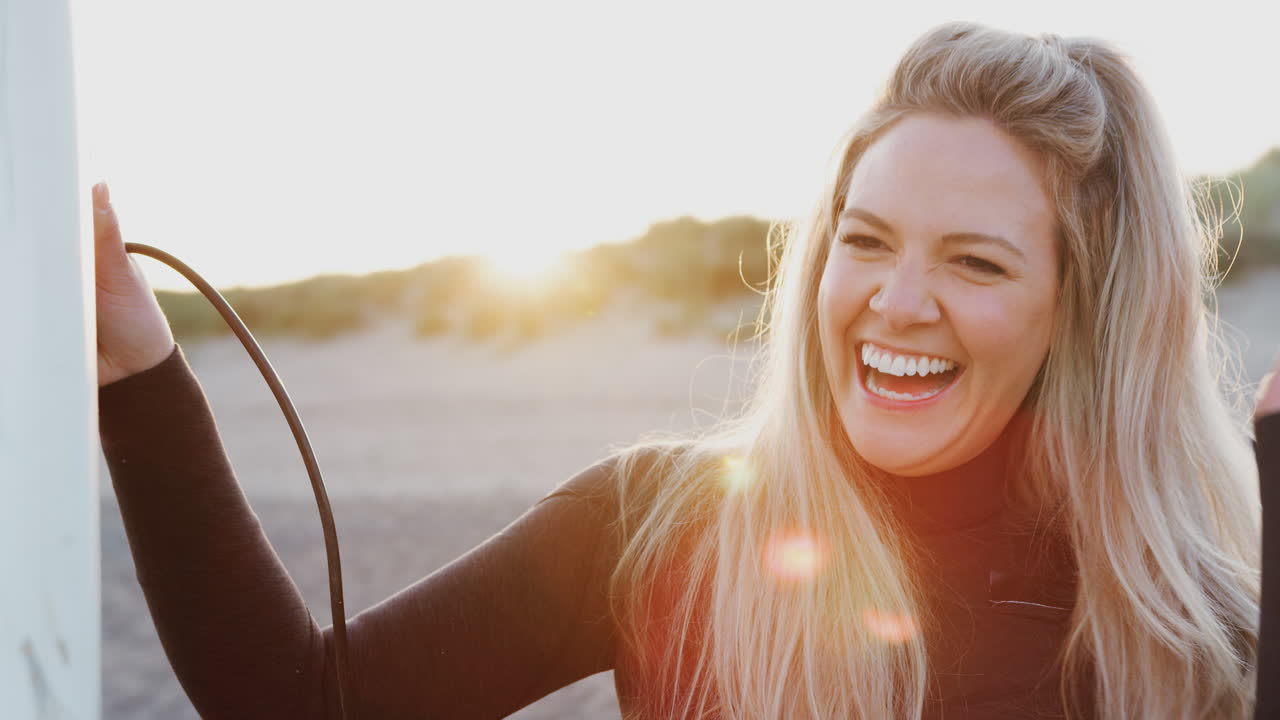 Woman Wearing Wetsuit Holding Surfboard Enjoying Surfing Vacation On Beach As Sun Sets