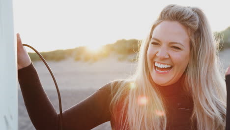frau mit wetsuit hält ein surfbrett und genießt den surfurlaub am strand, während die sonne untergeht