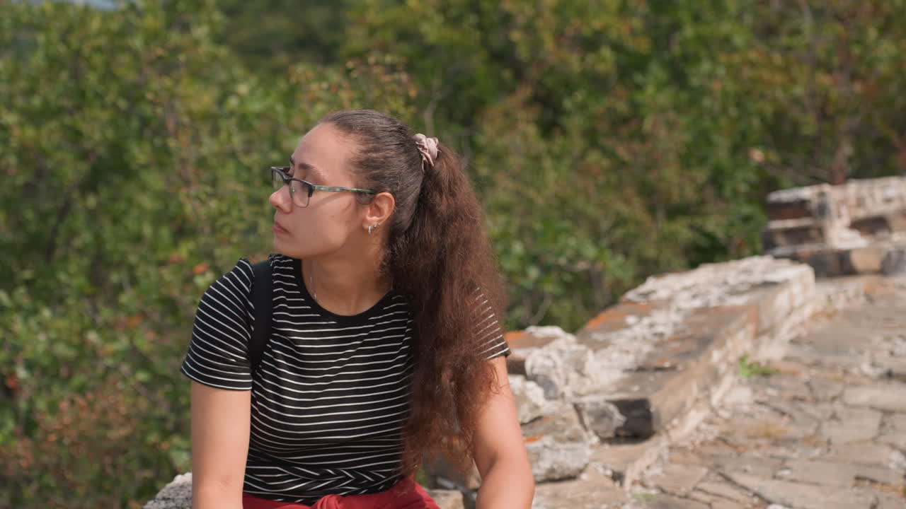 Giovane donna che riposa su un muretto lungo il sentiero con vista sulle montagne, riparandosi gli occhi con una mano, zaino da trekking e camicia a righe, sentiero in pietra che si snoda in lontananza, luce estiva, sguardo avventuroso, pausa turistica.
