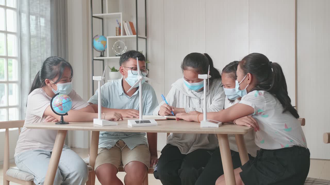 Middle School Classroom: Asian Kids Are Wearing Facemasks Learning About Ecology Problems And Their Solution With Wind Turbine Model And Globe At School