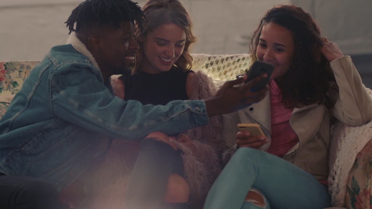 group of friends relaxing on rooftop at night sitting on couch young african american man using smartphone showing beautiful women social media lifestyle enjoying weekend