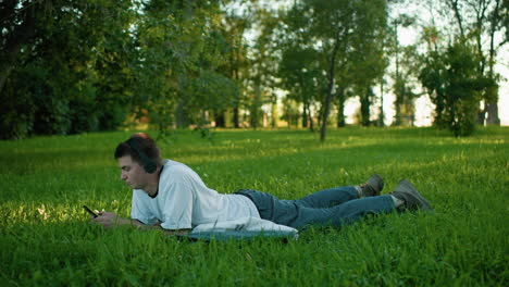 side view of young man lying on grassy field with headphones on, focused on his phone in a serene park surrounded by lush greenery, enjoying a peaceful outdoor moment