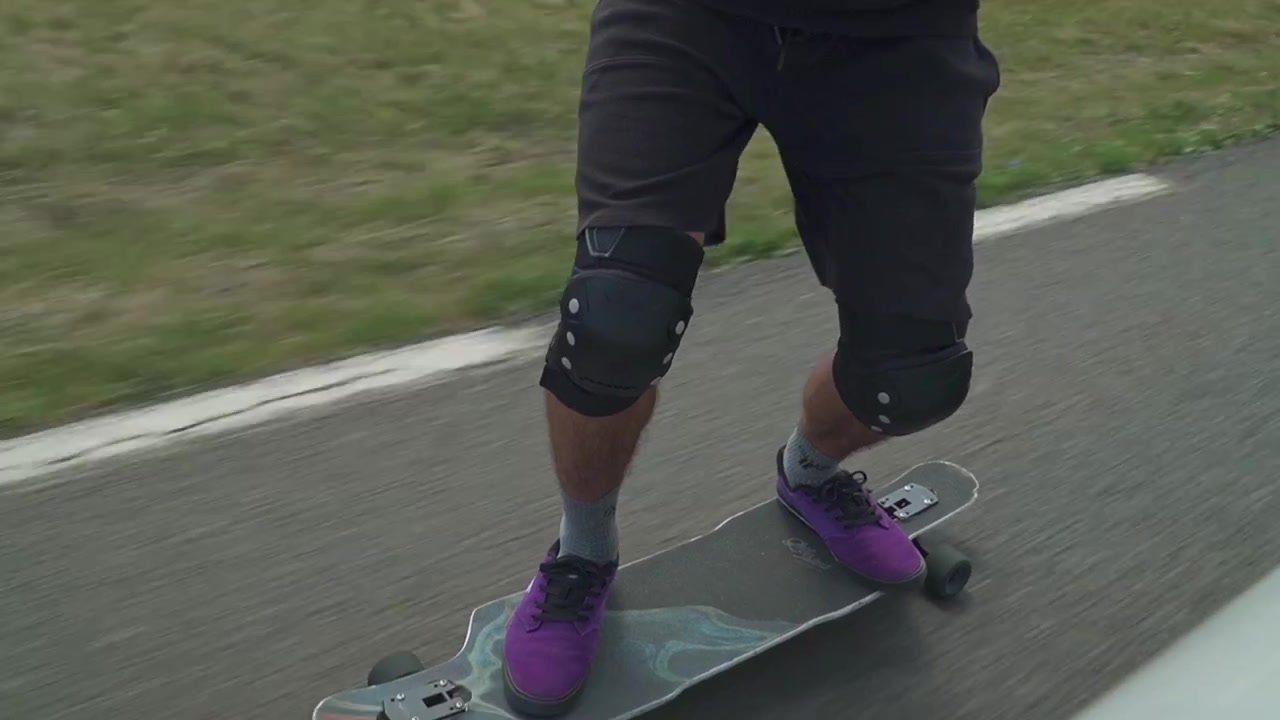 A skater in protective gear holds hands with a passenger in a moving car under cloudy skies.