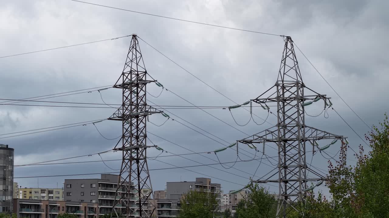 Two high-voltage transmission pylons and overhead cables above city apartment blocks under a cloudy sky, showing urban electricity infrastructure and utility corridor