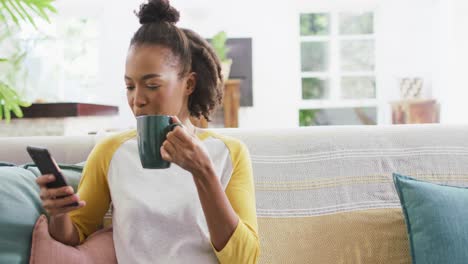 Mujer-Afroamericana-Tomando-Café-Y-Usando-Un-Teléfono-Inteligente-Sentada-En-El-Sofá-De-Casa