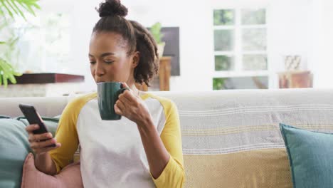 african american woman drinking coffee and using smartphone sitting on the couch at home