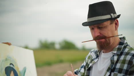 close-up of a sketcher wearing a hat and checkered shirt, deeply engrossed in painting. he holds a brush in his mouth while working on his artwork against a serene, grassy background