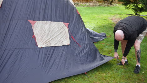 Ein-Mann,-Der-Bei-Regen-Auf-Einem-Campingplatz-Beim-Camping-Ein-Zelt-Abbaut-Und-Heringe-Entfernt