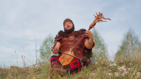 Performer-in-ethnic-clothes-plays-music-sitting-on-grass