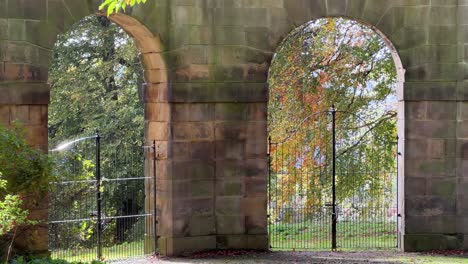 Alte-Steinbögen-Mit-Eisentoren-Und-Das-Sonnenlicht,-Das-Durch-Sie-Scheint,-Und-Goldene-Herbstbäume-In-Der-Ferne