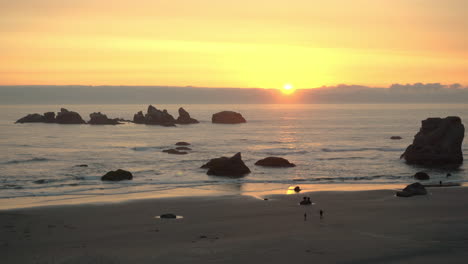 Puesta-De-Sol-Sobre-El-Océano-Pacífico-En-Bandon-Oregon-Con-Formaciones-Rocosas-De-Gatos-Y-Gatitos-Siluetas,-Gente-Irreconocible-Caminando-En-La-Playa