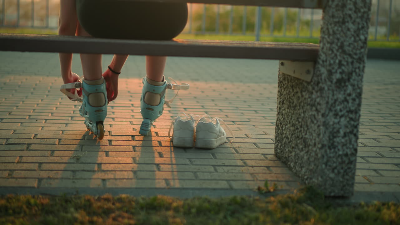 따뜻한 저녁 빛에 벤치에 앉아있는 젊은 여자의 뒷면은 왼쪽 롤러 스케이트의 줄을 제거하고, 바닥에 그녀의 에 두 개의  스니커를 가지고 있습니다.