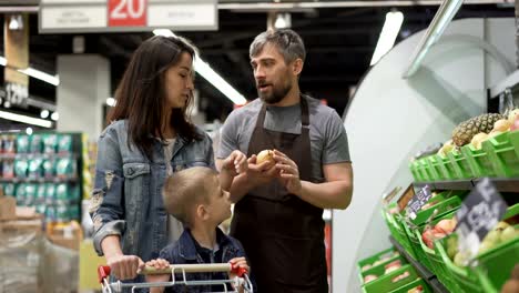 experienced shop assistant is selling fruit to cheerful young family mom and child, giving them pears, apples and pineapple, happy customer is laughing and smiling.