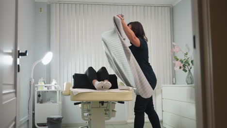 massage therapist spreads gray cloth over female client on cream colored massage chair in bright white treatment room lit by lamp, creating hygienic, comfortable setup for upcoming wellness session