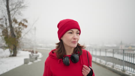 close-up of woman with headphones around neck walking along interlocked pathway with bag over shoulder, subtle smile, iron railing nearby, and blurred cityscape view in foggy atmosphere