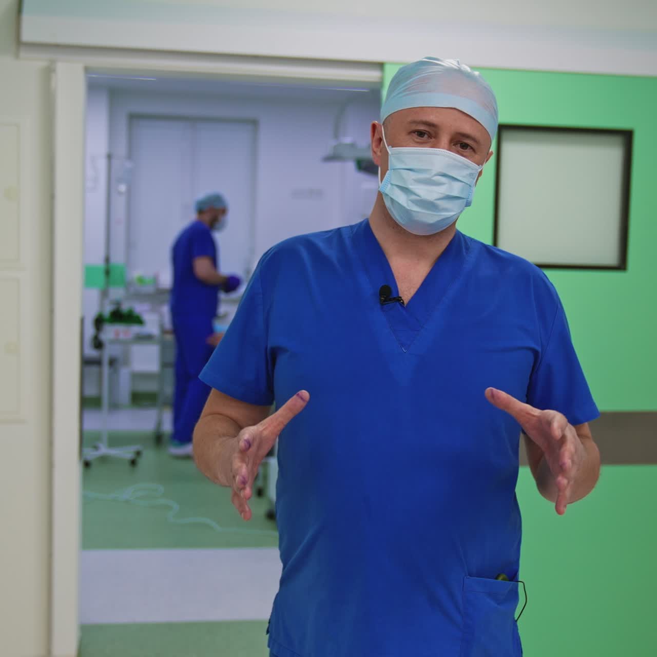 Plastic surgeon talking on camera. Portrait of a male doctor in blue medical uniform and mask speaking on the background of an operating room in clinic