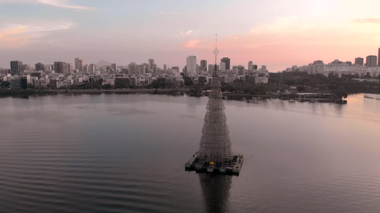 Veduta paesaggistica con l'albero di Natale galleggiante più alto del mondo del 2018 a Rio de Janeiro, sul lago cittadino, arricchita da un tramonto incredibilmente colorato, con una panoramica verso l'alto che svela il quartiere di Ipanema sullo sfondo.