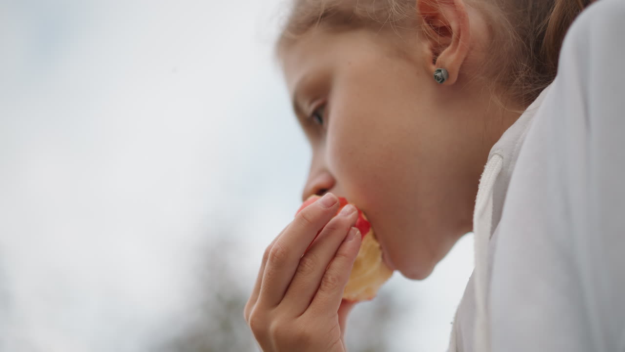 Young girl eating apple outdoors, closeup portrait, soft overcast sky, white jacket, tiny earring, slow contemplative bite, sticky juice on fingers, childhood innocence, peaceful afternoon snack