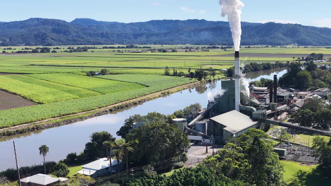Aerial footage captures a sugar mill emitting steam amidst lush green fields and a winding river under clear skies