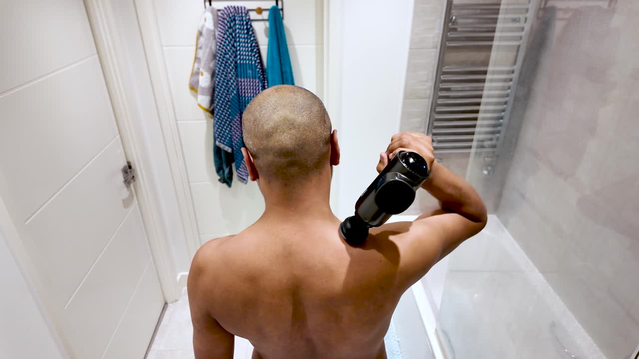 Shirtless man massaging his back with percussion massage gun in bathroom. High Angle, Slow Motion