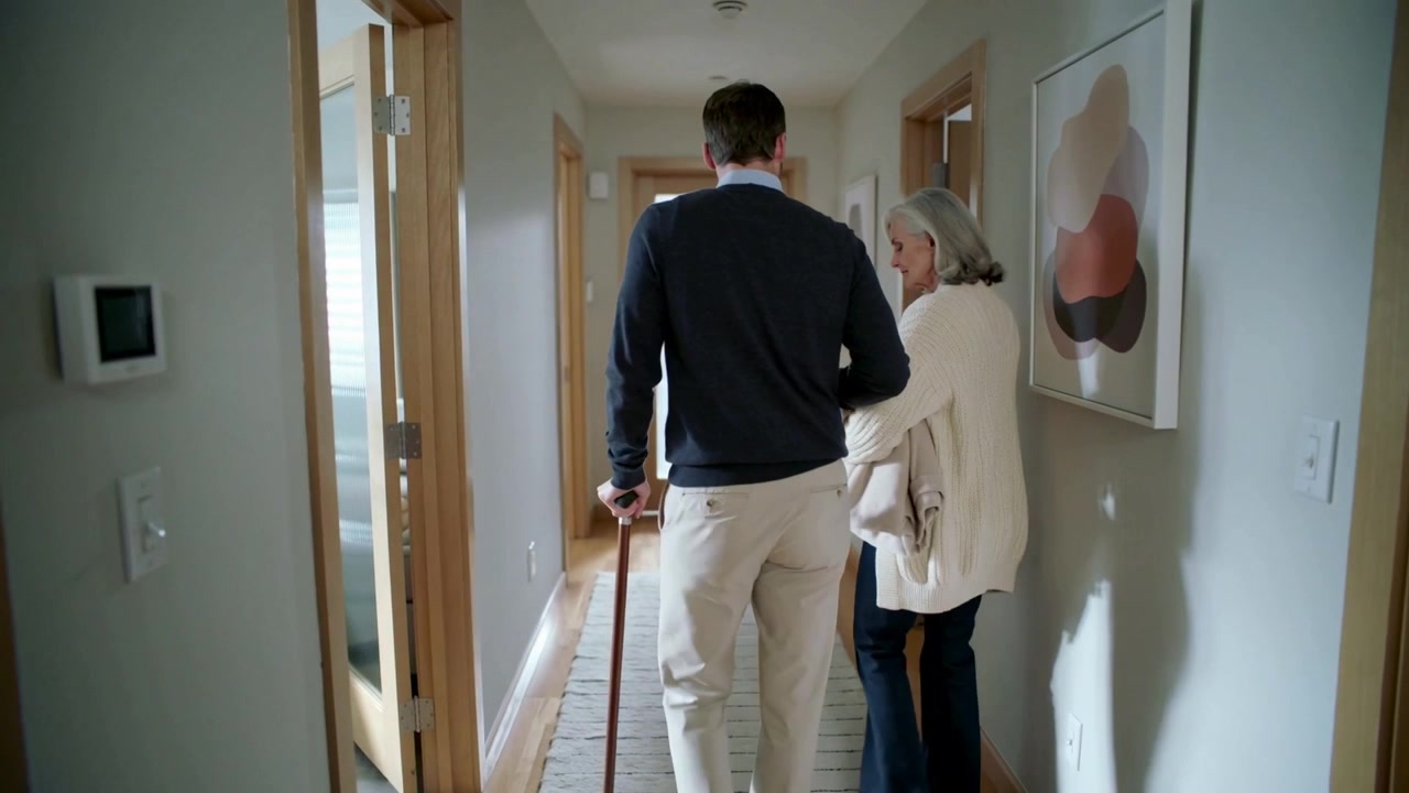Steadying on wooden cane, adult male in navy sweater guiding senior female down runner toward light
