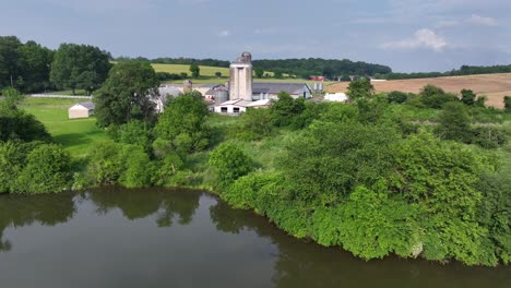 aerial view showing growing farm plantation fields in front of large farmstead with silo. drone shot on cloudy day in pennsylvanian countryside. river in foreground