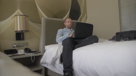 a middle-aged white man is sitting or lying slouched in the bed of his modern hotel room while working on his laptop