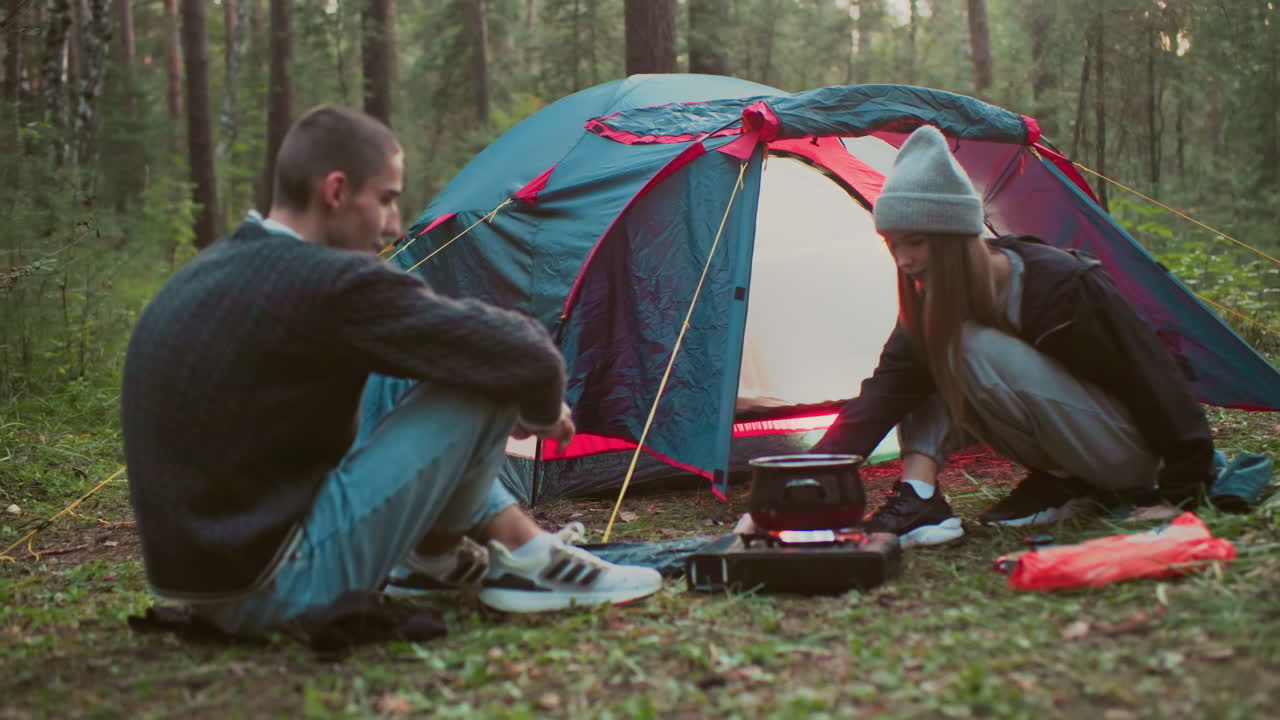 young couple cooking outdoors as woman prepares food on portable stove near tent while man sits on ground watching surrounded by forest trees camping gear