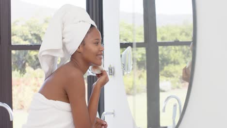 Happy-biracial-woman-with-towel-on-head-looking-in-mirror-washing-her-face,-slow-motion