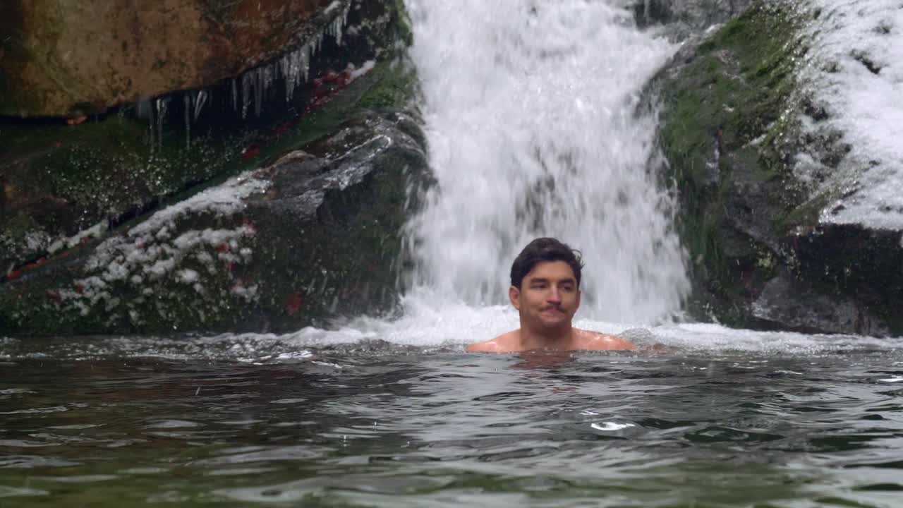 hombre guapo nadando en el hielo en un río frío bajo cero junto a la hermosa cascada en las montañas nevadas durante el día de invierno - tiro estático, cámara lenta