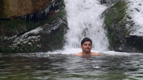 Hombre-Guapo-Nadando-En-El-Hielo-En-Un-Río-Frío-Bajo-Cero-Junto-A-La-Hermosa-Cascada-En-Las-Montañas-Nevadas-Durante-El-Día-De-Invierno---Tiro-Estático,-Cámara-Lenta