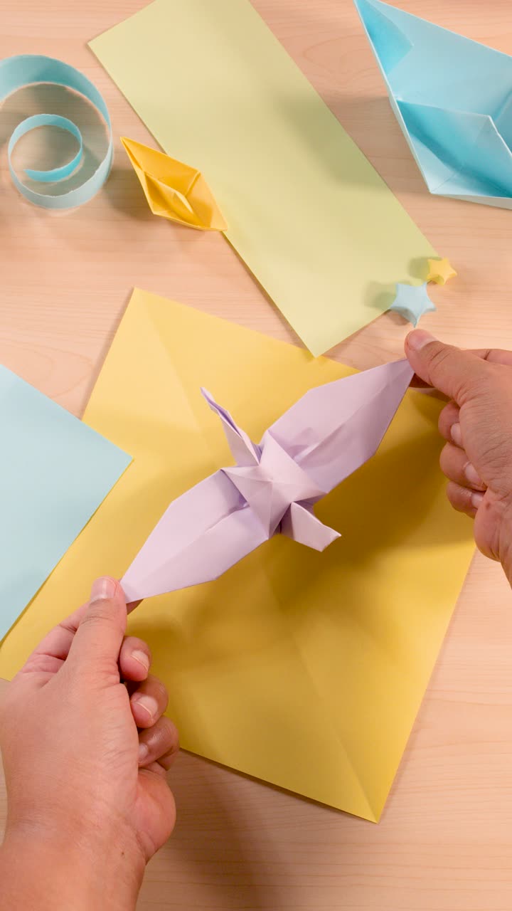 Person folds white origami bird with pastel paper sheets, overhead view, soft even lighting