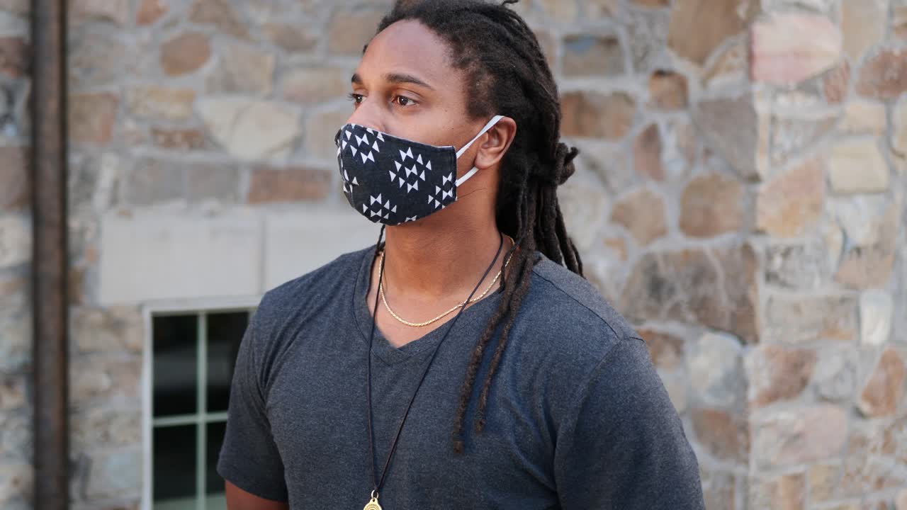 African American man with dreadlocks standing and waiting as he pulls his Covid-19 mask up and looks off
