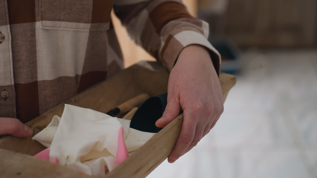 Female hands holding wooden container filled with towel and vintage wooden tool on bright tiled floor near modern interior with potted plants and soft natural light through glass window