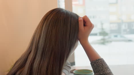 side view of student seated indoors supporting head with hand while looking out transparent cafeteria window during snowy day with phone on table and breakfast beside her as cars pass on the street