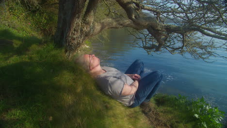 woman lying down asleep relaxing under a tree in warm sunlight next to lake waterfront - static