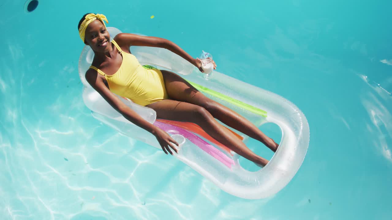 Attractive african american woman lying on inflatable holding drink and smiling in swimming pool