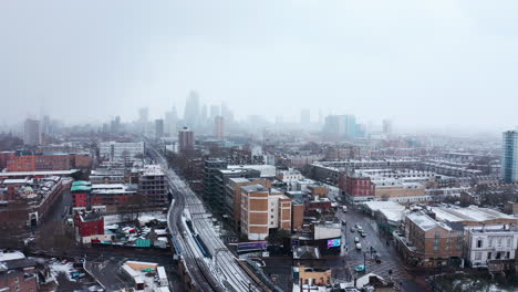 circling aerial shot of train tracks covered in show heading towards city of london skyscrapers