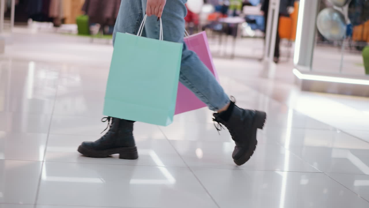 소매점에서 빛의 반사와 함께 우아한 타일에 쇼핑 가방을 가지고 는 사람의 측면 시각, 배경은 바쁜 쇼핑몰에서 옷을 매달리고 랙을 보여줍니다.