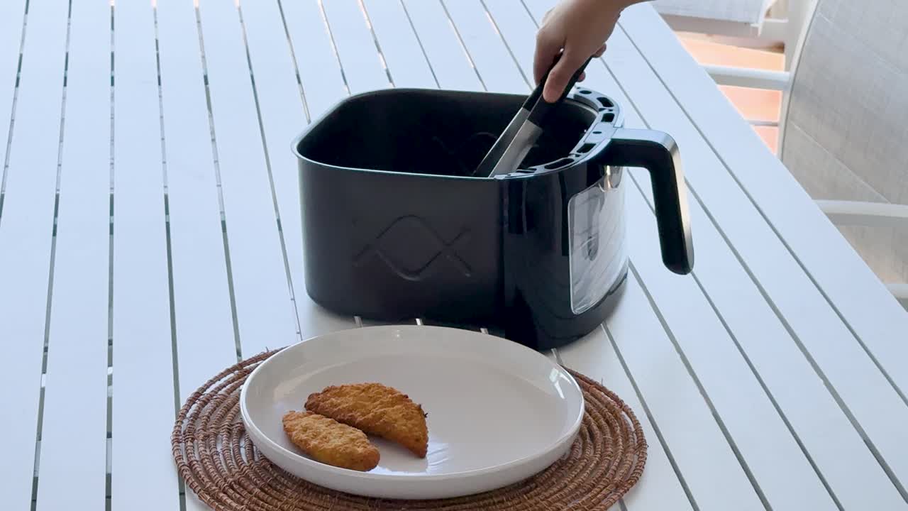 A hand uses tongs to transfer golden crumbed chicken tenders from a black air fryer basket onto a white plate in bright natural light