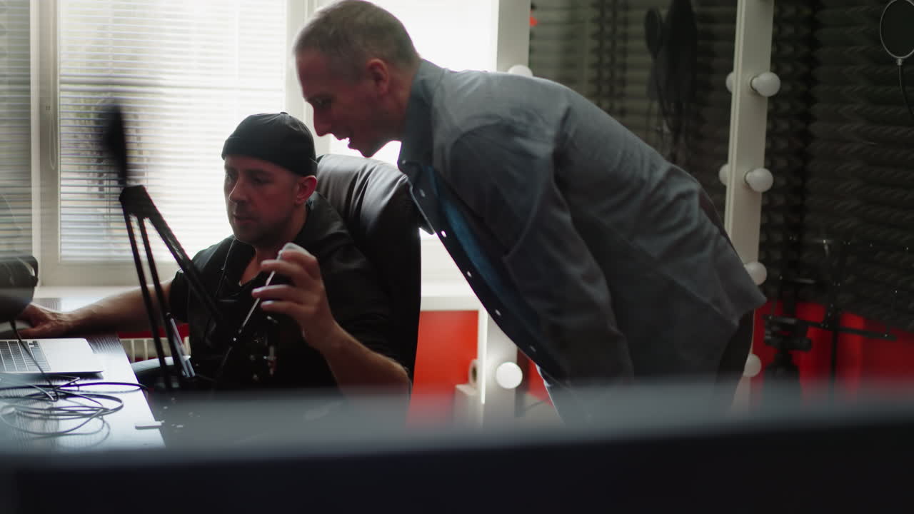 두 남자가 스튜디오 방에 앉아 있습니다. 한 명은 검은 옷을 입고 컴퓨터 앞에 앉은 프로듀서일 가능성이 높습니다. 다른 한 명은 긴 팔을 입은 셔츠를 입고 마이크를 테스트하기 위해 몸을 기울이고 있습니다.