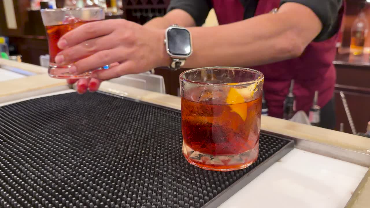 Bartender adds citrus garnish to cocktails, then serves drinks on bar mat under warm lighting