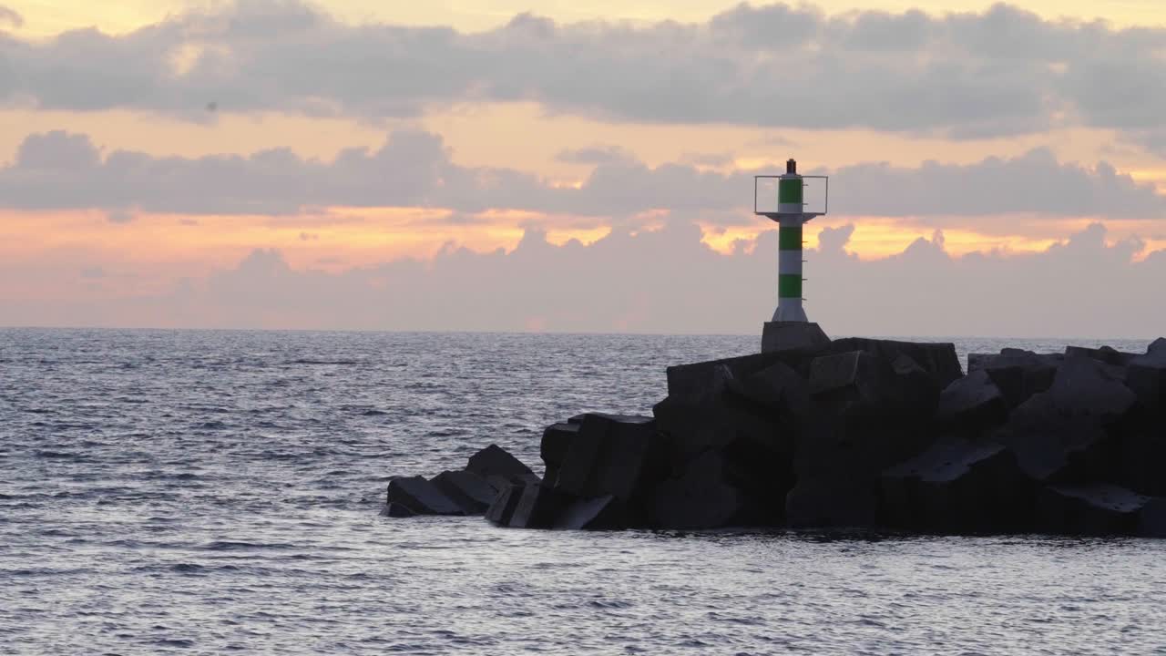 A lighthouse stands tall on rocky cliffs as the sun sets behind it, casting a golden glow on the sea. Waves crash gently against the shore, creating a peaceful and serene coastal scene.