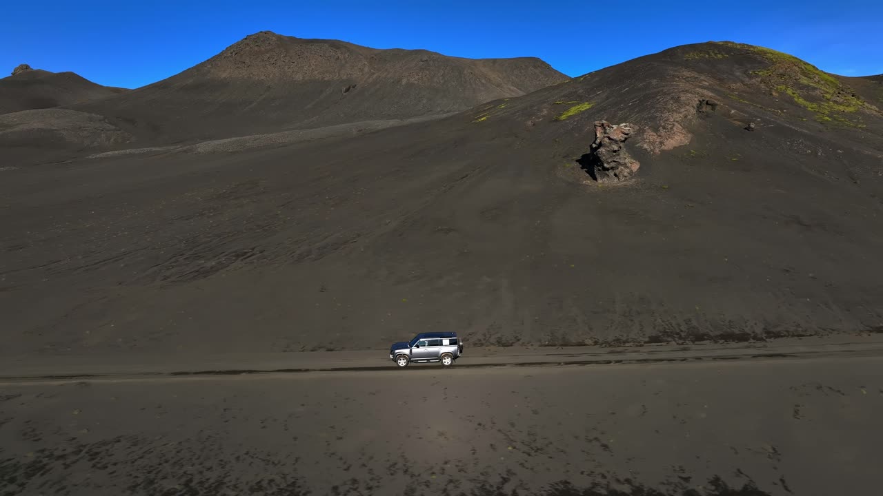 Vista laterale con drone di un SUV 4x4 che guida lungo un sentiero polveroso sotto scure colline vulcaniche con chiazze di muschio verde sotto un cielo azzurro e sereno. Scena di avventura fuoristrada isolata