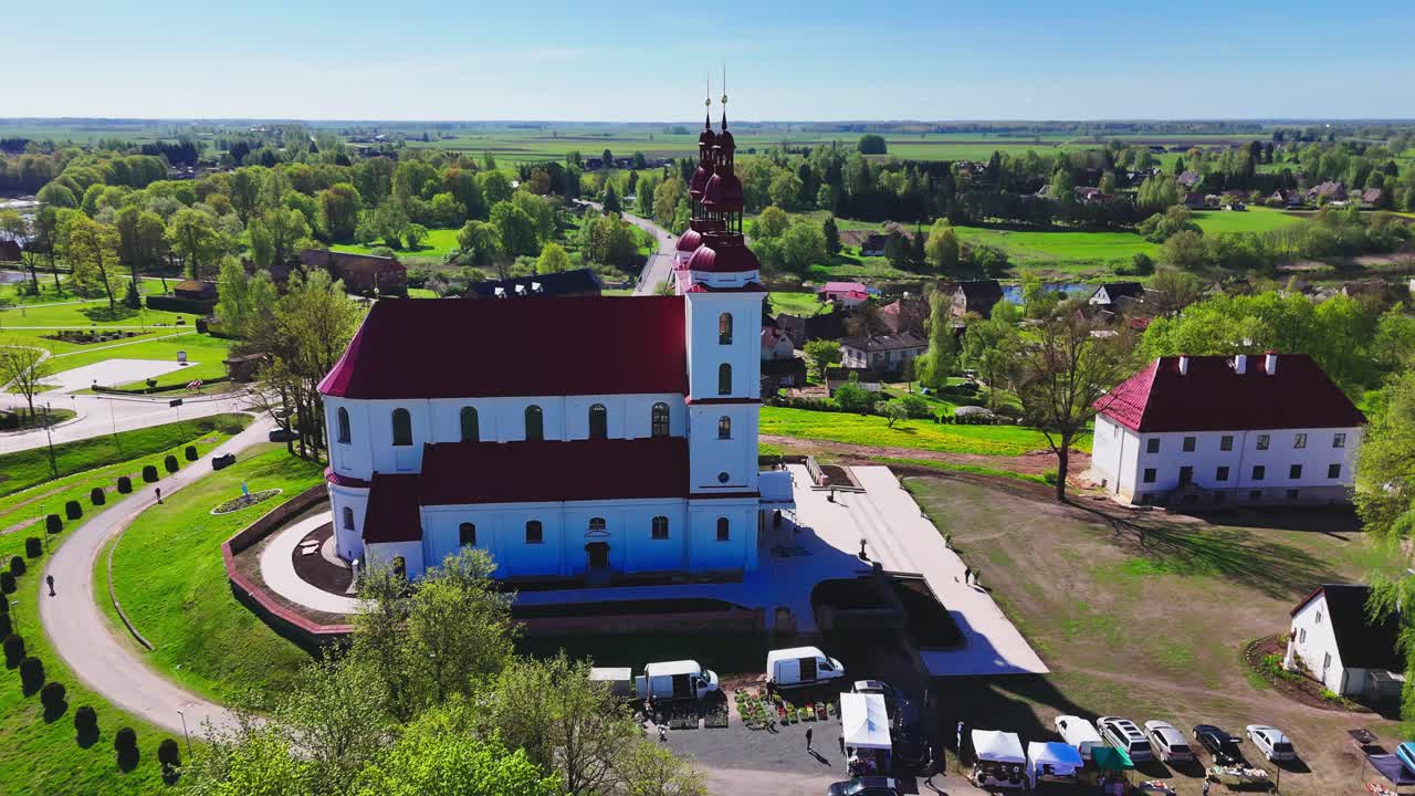 Aerial view of a historic twin-spired church in a European village, with red rooftops, farmland, winding roads, and a backdrop of lush green countryside under clear skies.