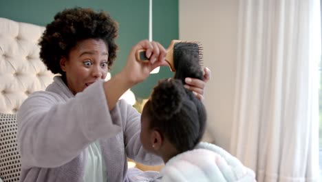 happy african american mother combing daughter hair sitting on bed in bedroom, slow motion