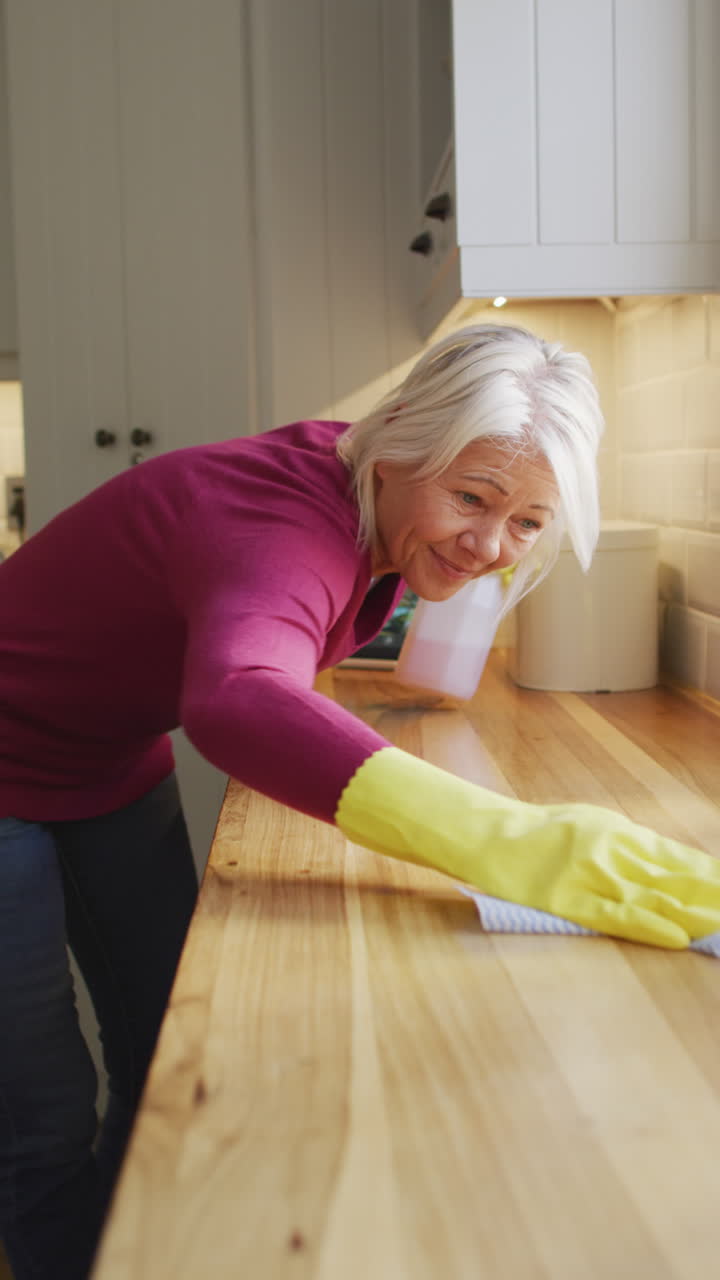 una mujer caucásica senior feliz con guantes de goma limpia la encimera de la cocina en video vertical