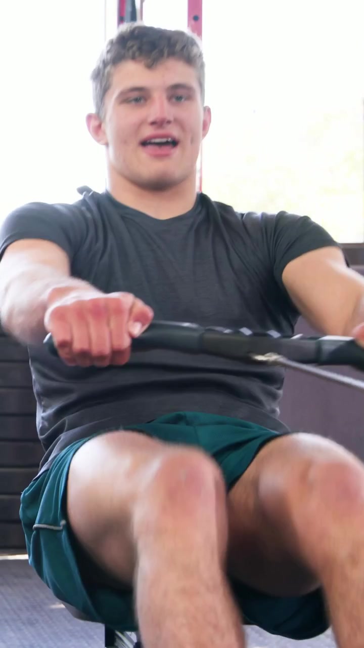 Video verticale: Uomo che rema con maglietta grigio scuro e pantaloncini verde acqua, tira la maniglia su un vogatore da palestra.