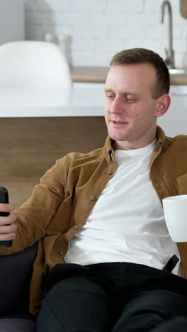 handsome man using a phone at home. guy rests on sofa with a cup of coffee and doing photos on a smart phone in the kitchen. vertical video