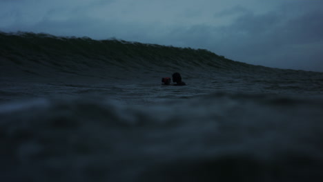 surfers wait in water as ocean wave rises above and crashes into barrel of green and dark water